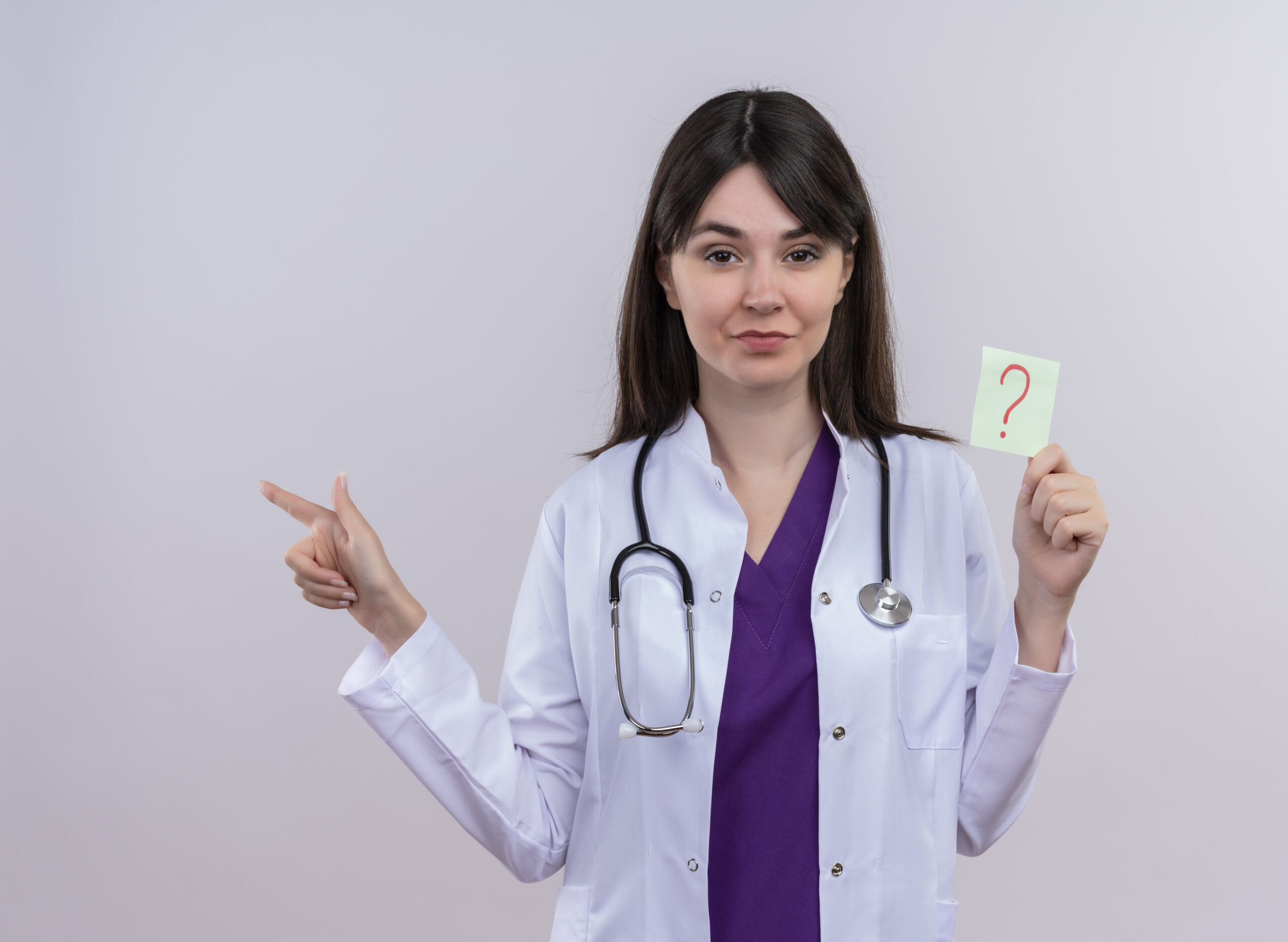 confident young female doctor in medical robe with stethoscope points to the side and holds question mark on isolated white background with copy space