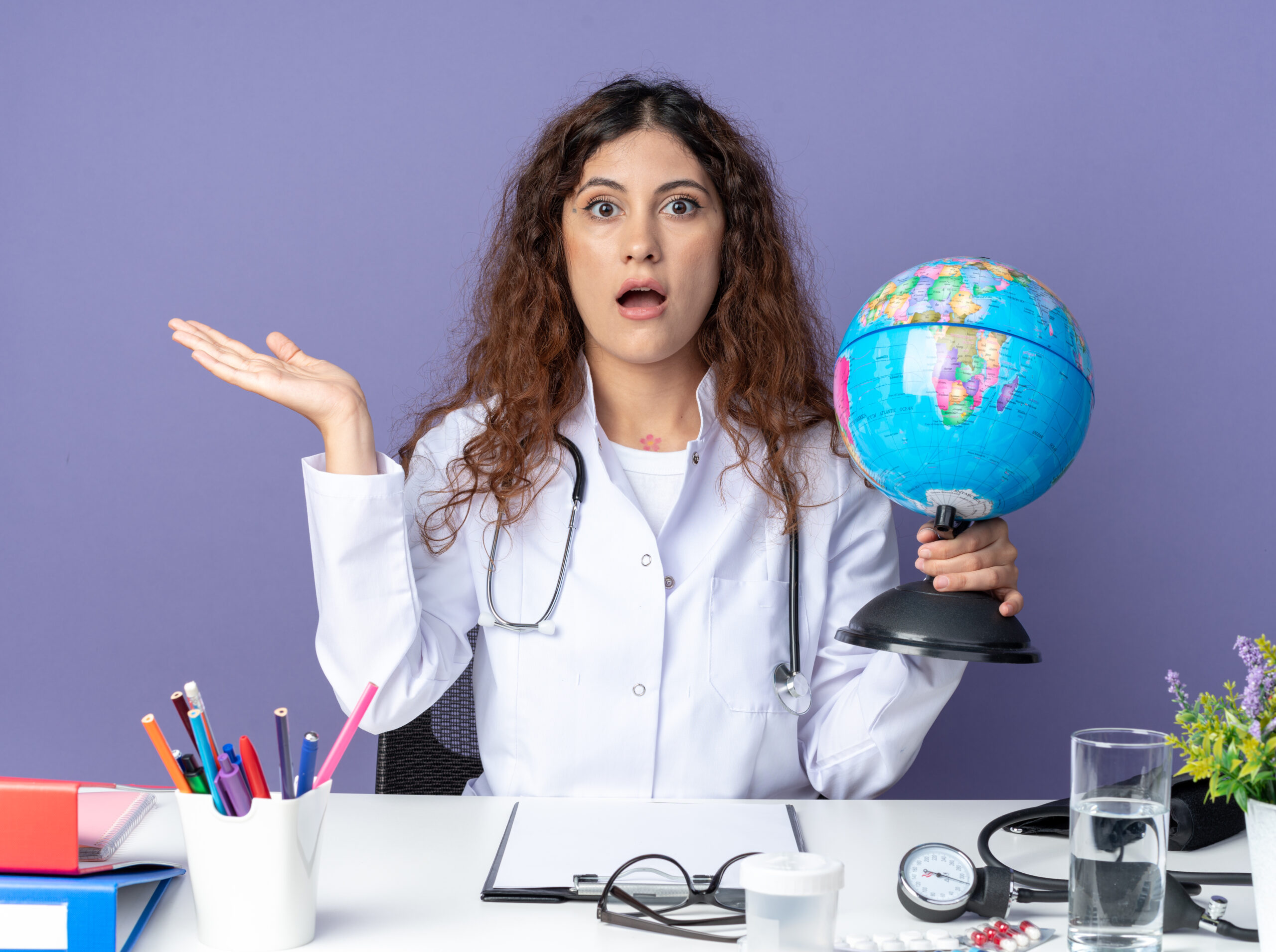impressed young female doctor wearing medical robe stethoscope sitting table with medical tools holding globe looking front showing empty hand isolated purple wall